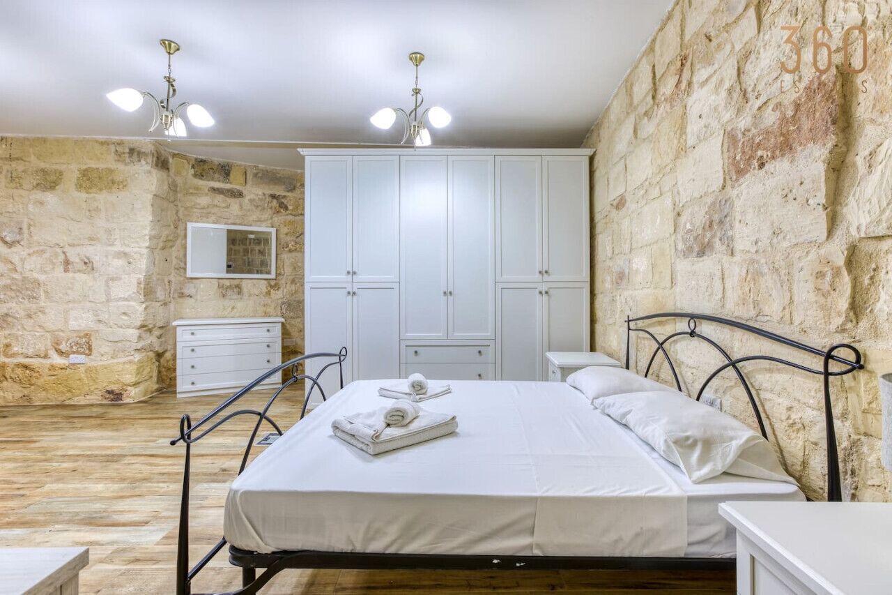 Una camera da letto con parete in pietra, un letto con lenzuola bianche, un armadio e un comodino.