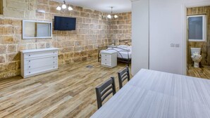 A room with a stone wall, a bed, a bedside table, a dresser, a TV mounted on the wall, and a window. - Lovely 1BR Studio home in Quiet Town of Zejtun (Iż-Żejtun)