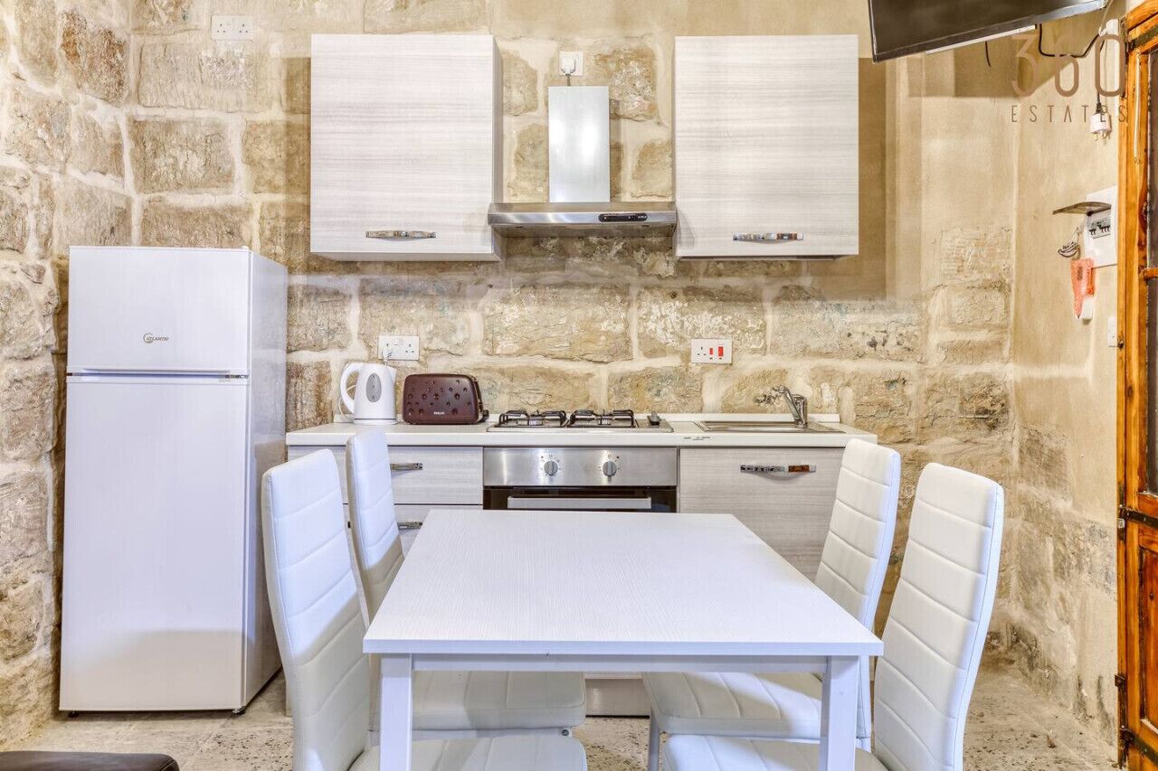 Una cucina compatta con frigorifero bianco, piano cottura a gas e forno a microonde. È presente un tavolo da pranzo con sedie bianche.