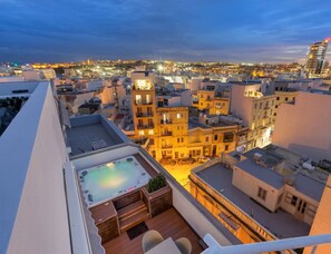 A rooftop terrace with a hot tub, a pool, and a view of the cityscape at dusk.