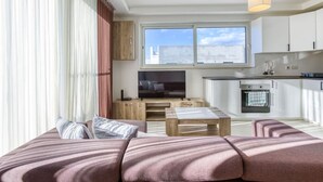 A modern living room with a large sofa, a coffee table, and a TV. There is a kitchen area with cabinets and a window. - Sunny 2BR Penthouse with Spacious Rooftop Terrace (San Ġwann)