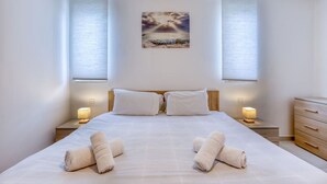 A neatly made bed with two pillows and two rolled towels, a wooden nightstand with a lamp, a wooden dresser, and a painting of a sunset over the ocean on the wall. - Sunny 2BR Penthouse with Spacious Rooftop Terrace (San Ġwann)
