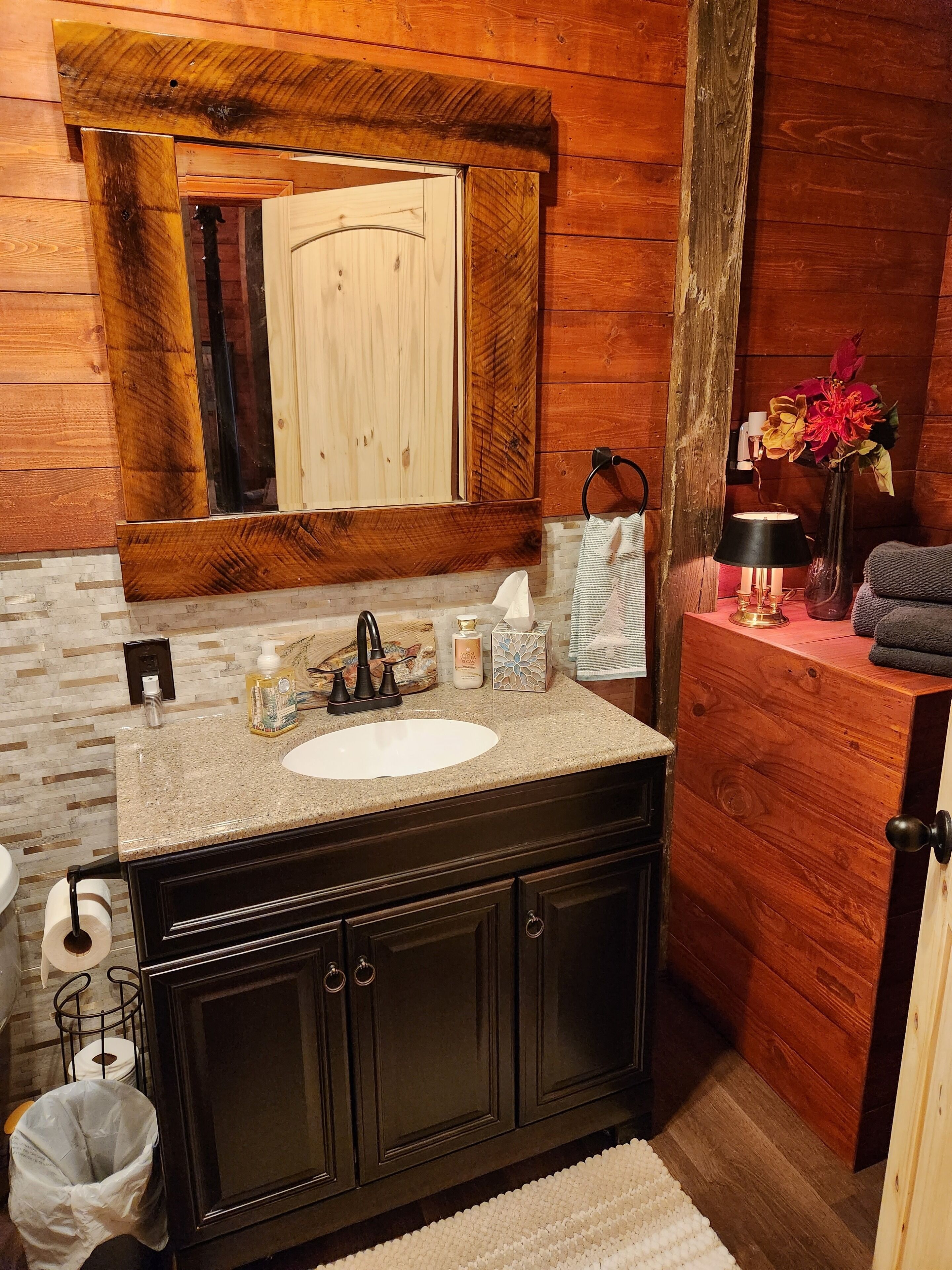 A wooden cabin bathroom with a granite sink, dark cabinets, a mirror, and a small bedside table with a lamp and flowers.