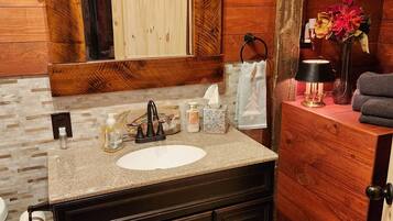 A wooden cabin bathroom with a granite sink, dark cabinets, a mirror, and a small bedside table with a lamp and flowers.