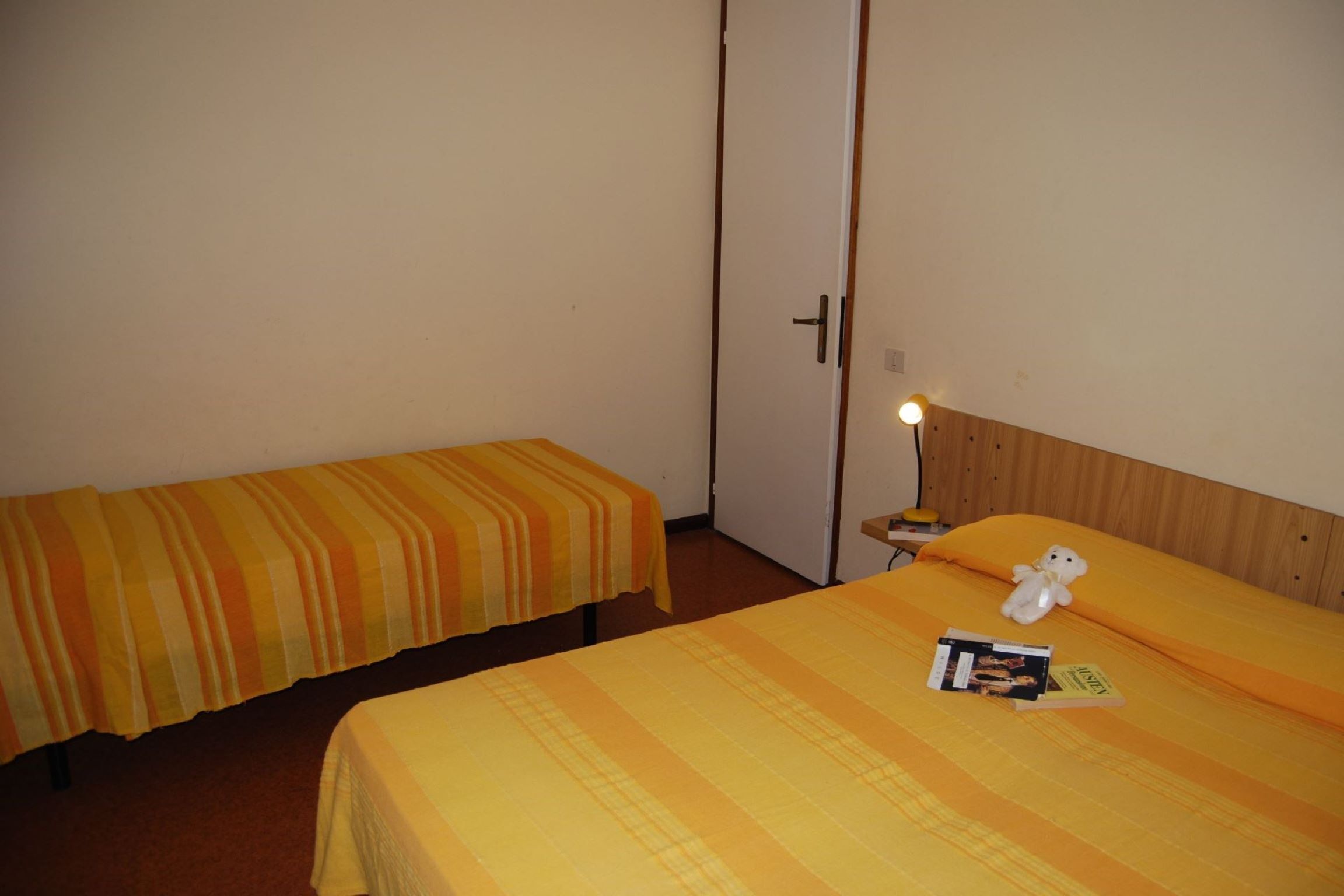 A hotel room with two single beds, each with a striped blanket, a bedside table with a lamp, and a stuffed toy on one bed.