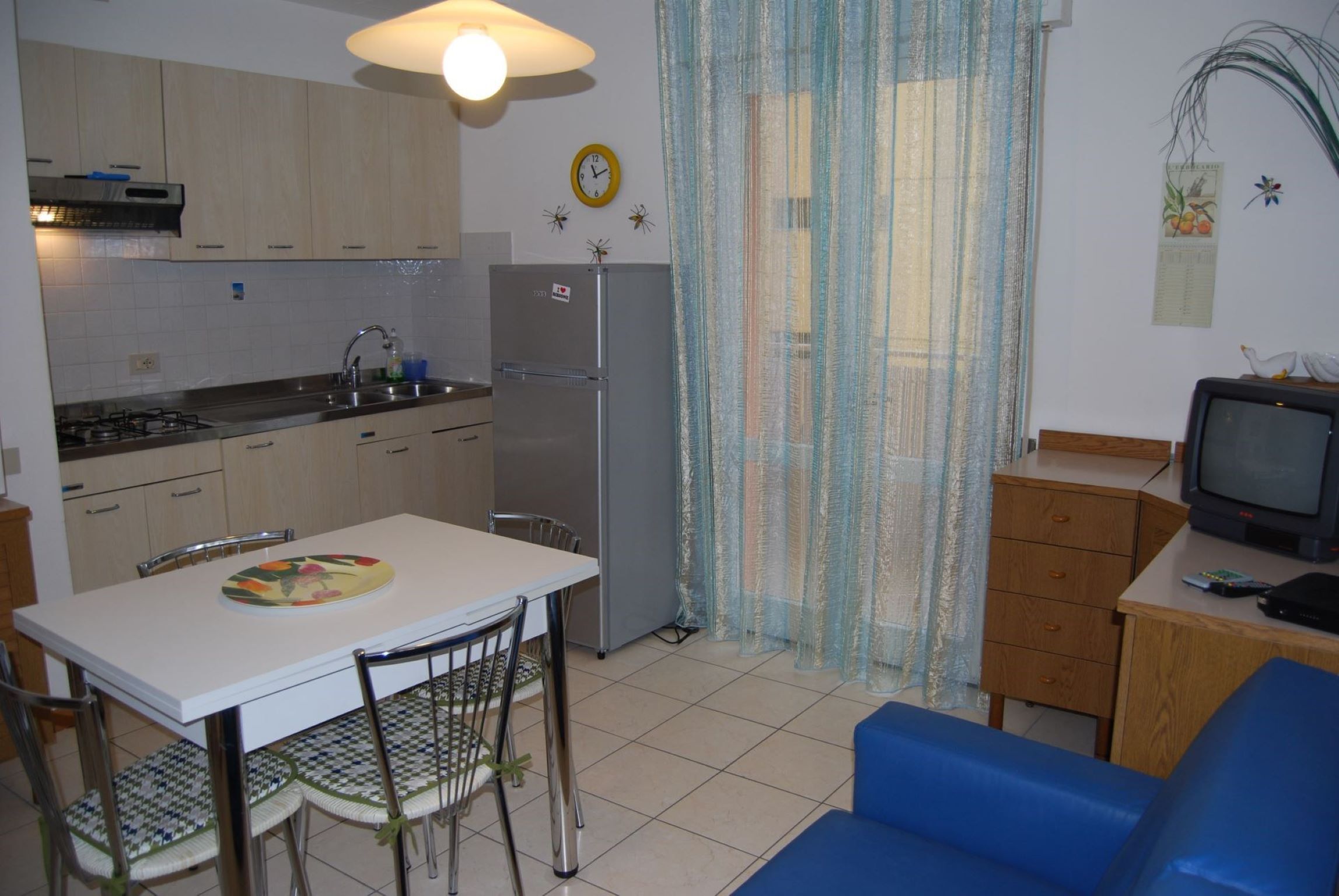 A compact kitchen with a refrigerator, stove, and sink. There's a dining table with chairs and a blue sofa. A television is on a wooden table.