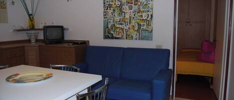 A room with a blue sofa, a white table with a placemat, a television, and a colorful abstract painting on the wall.