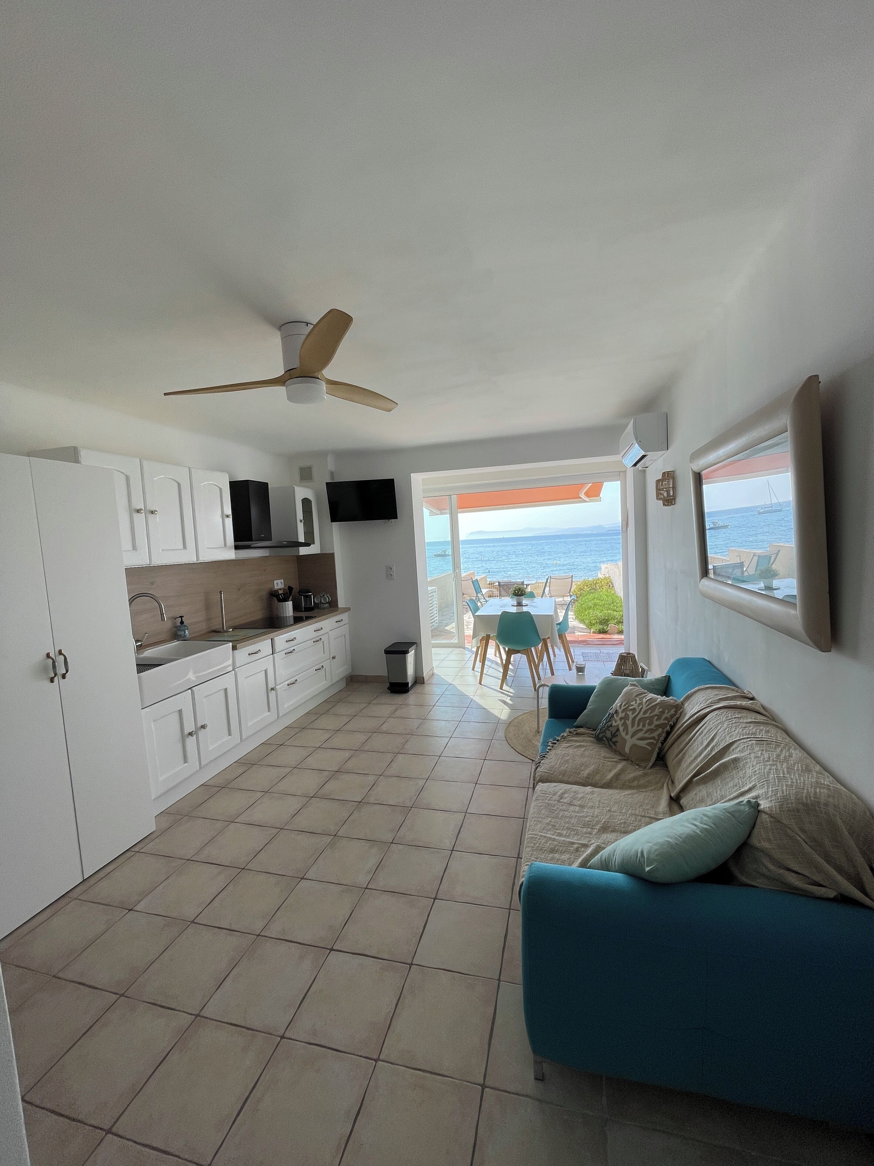 A modern kitchen with white cabinets, a ceiling fan, and a view of the sea through an open door.