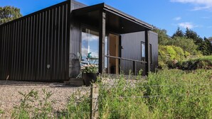 A modern, dark-colored building with a covered outdoor area, a gravel driveway, and a metal fence. - Core-na-bye (Inverness)