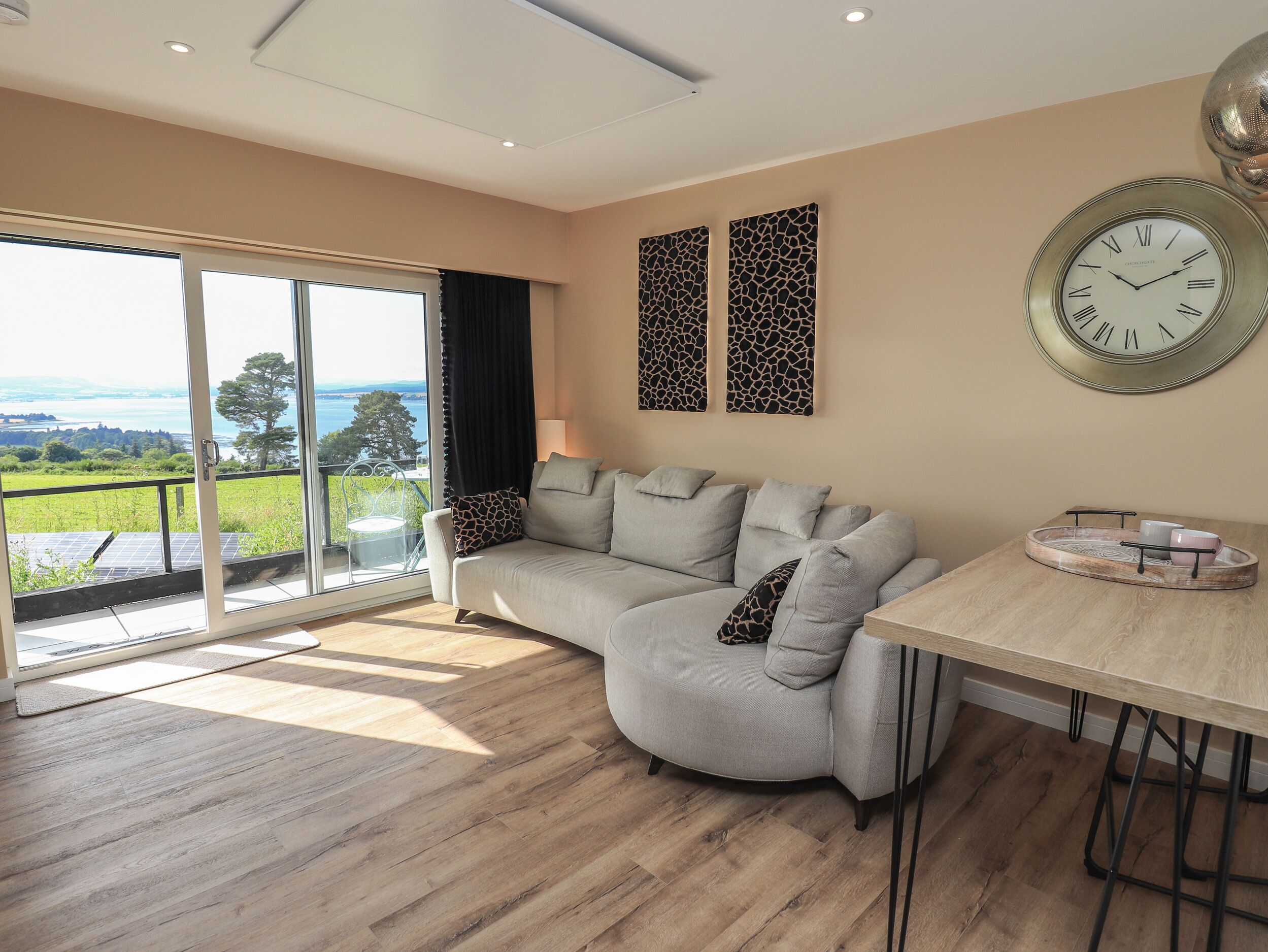 A modern living room with a grey sofa, a dining table with a tray, and a wall clock.