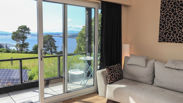 A room with a sofa, a table with a glass top, and a view of a lake through a sliding glass door. - Core-na-bye (Inverness)