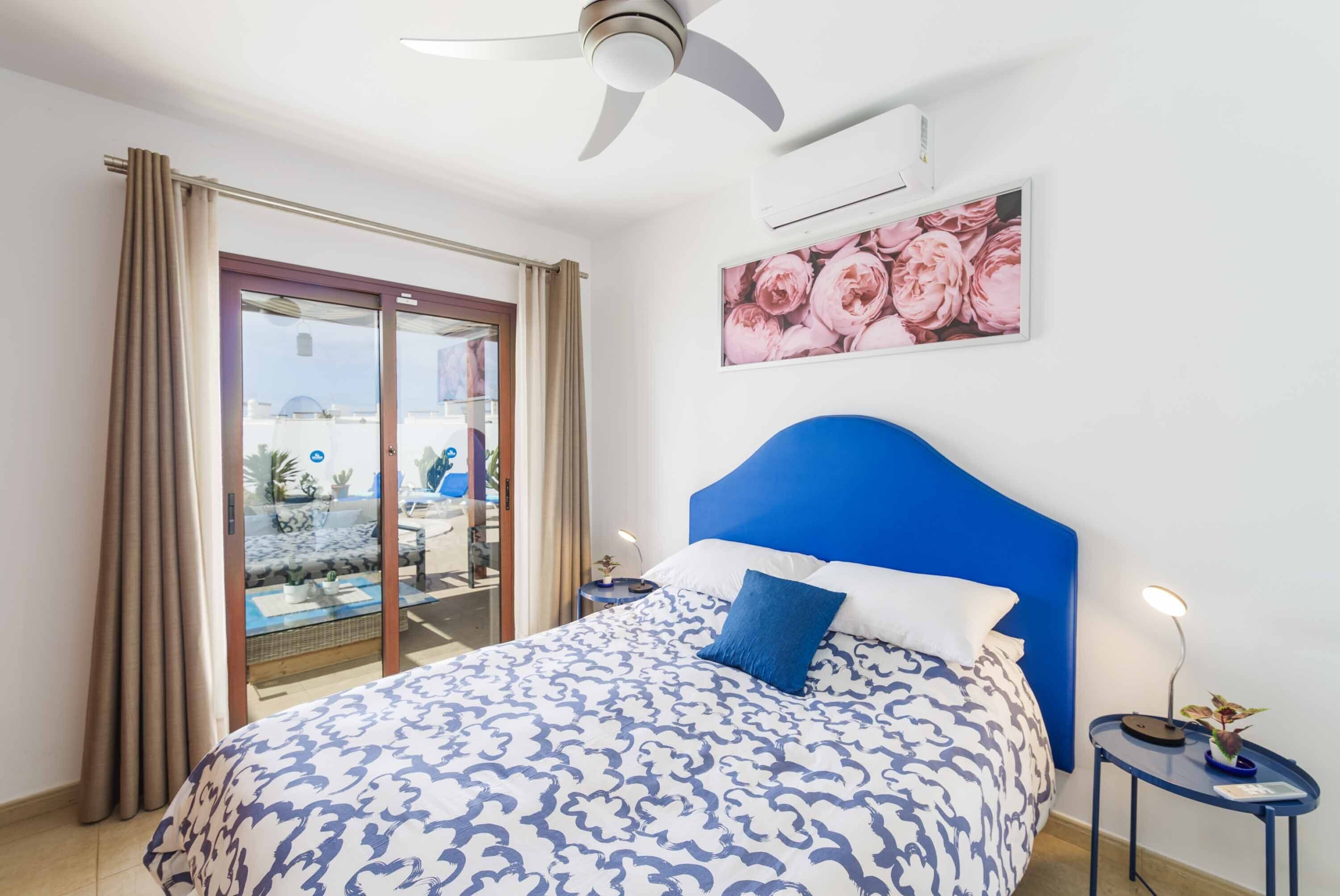 Une chambre à coucher avec un lit, un ventilateur de plafond, une table de chevet avec une lampe, un tableau représentant des fleurs et une porte-fenêtre coulissante donnant sur l’extérieur.