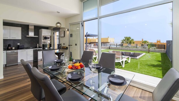 Una cocina moderna con electrodomésticos de acero inoxidable, un comedor con mesa y sillas de vidrio, y vistas a un césped verde y edificios.