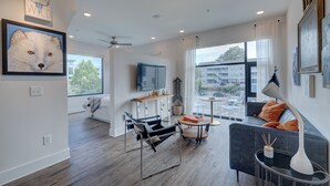 A modern living room with a sofa, a coffee table, and a chair. There is a TV mounted on the wall, a ceiling fan, and a window with a view of buildings outside.