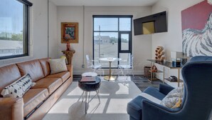 A modern living room with a leather sofa, a round coffee table, and a blue armchair. There is a large window with a view of buildings outside. - The Hive 203 Sweeter by BlueStar (Wilmington)