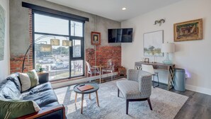 A modern living room with a large window, a sofa, a patterned armchair, a small table, and a desk with a lamp. - The Hive 206 Sweeter (Wilmington)