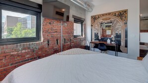 A bedroom with a brick wall, a large window, a bed, a desk, and a decorative mirror. - The Hive 207 Bees Knees by BlueStar (Wilmington)