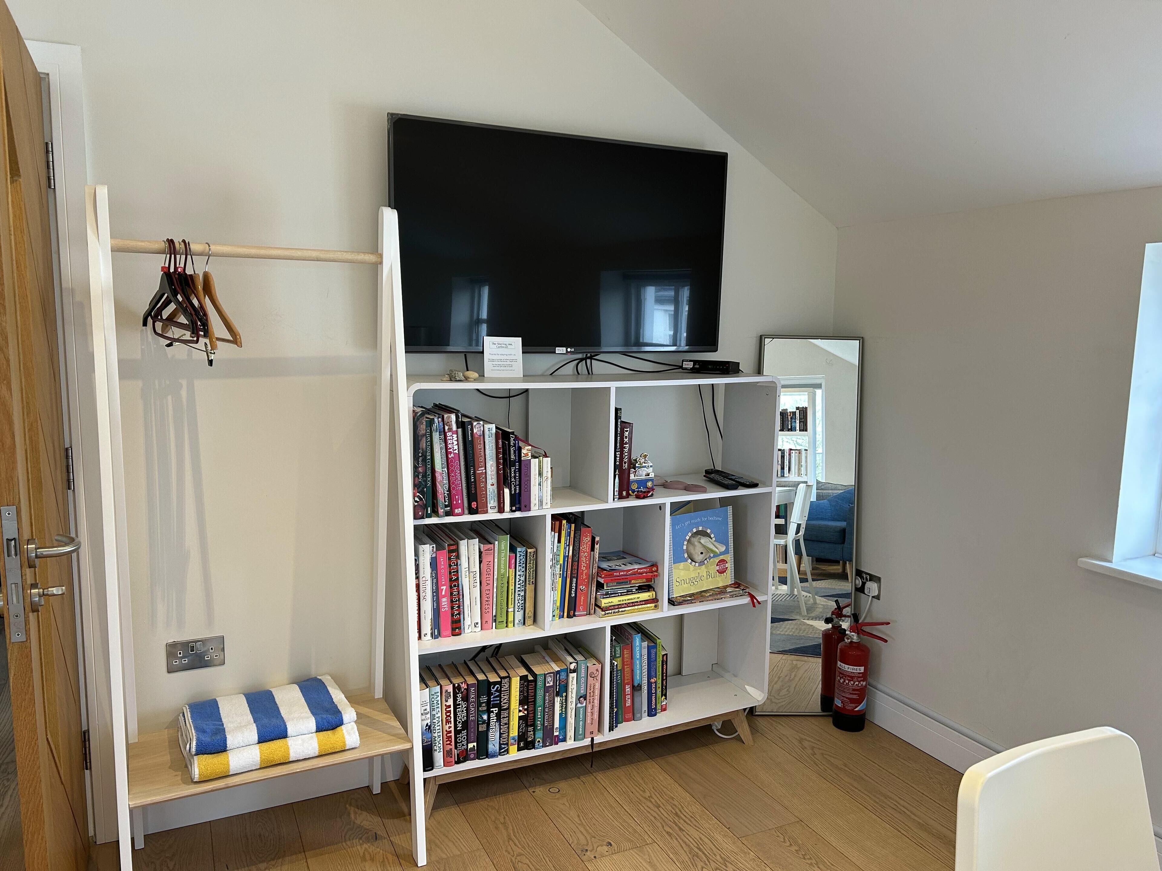 Una habitación con una estantería blanca llena de libros, un televisor de pantalla plana montado arriba, un extintor y una ventana con vistas al exterior.