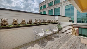 Un patio in legno con due sedie bianche e un tavolino, una fila di sculture in pietra e un edificio con ampie finestre sullo sfondo.