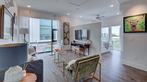 Un soggiorno moderno con un divano, una poltrona verde, un tavolino da caffè e una TV. È presente un'ampia finestra con vista sugli edifici.