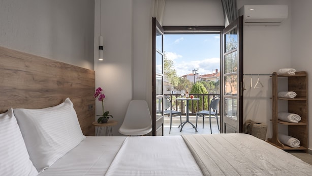 A modern hotel room with a wooden headboard, a large bed with white linens, a small table with a potted plant, a white chair, and a balcony with a table and chairs. - Eleon Suites & Apartments (Kassandra)
