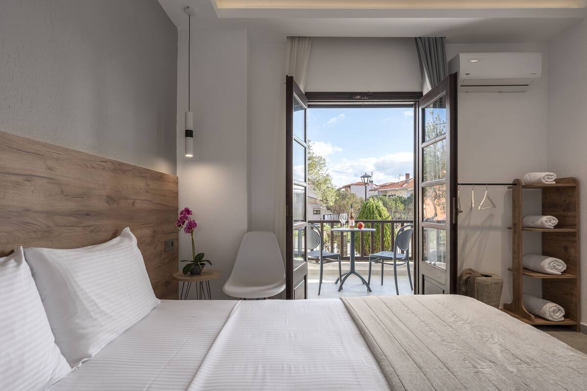 A modern hotel room with a wooden headboard, a large bed with white linens, a small table with a potted plant, a white chair, and a balcony with a table and chairs.
