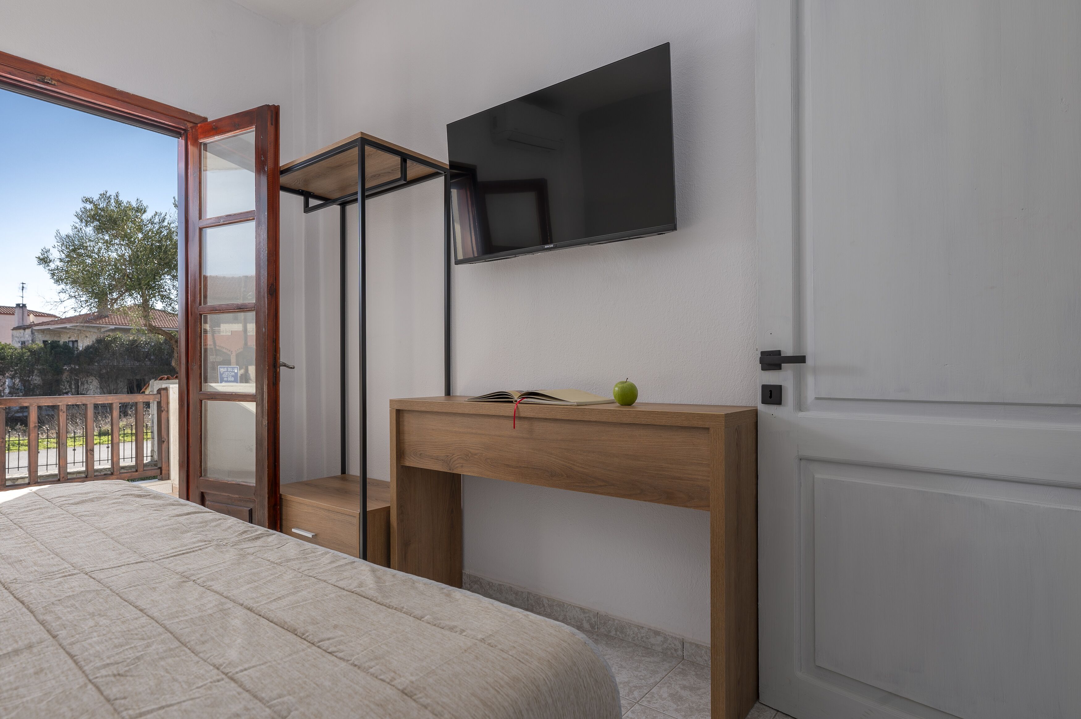 A bedroom with a bed, a wooden desk, a TV mounted on the wall, and a balcony with a view of the outside.