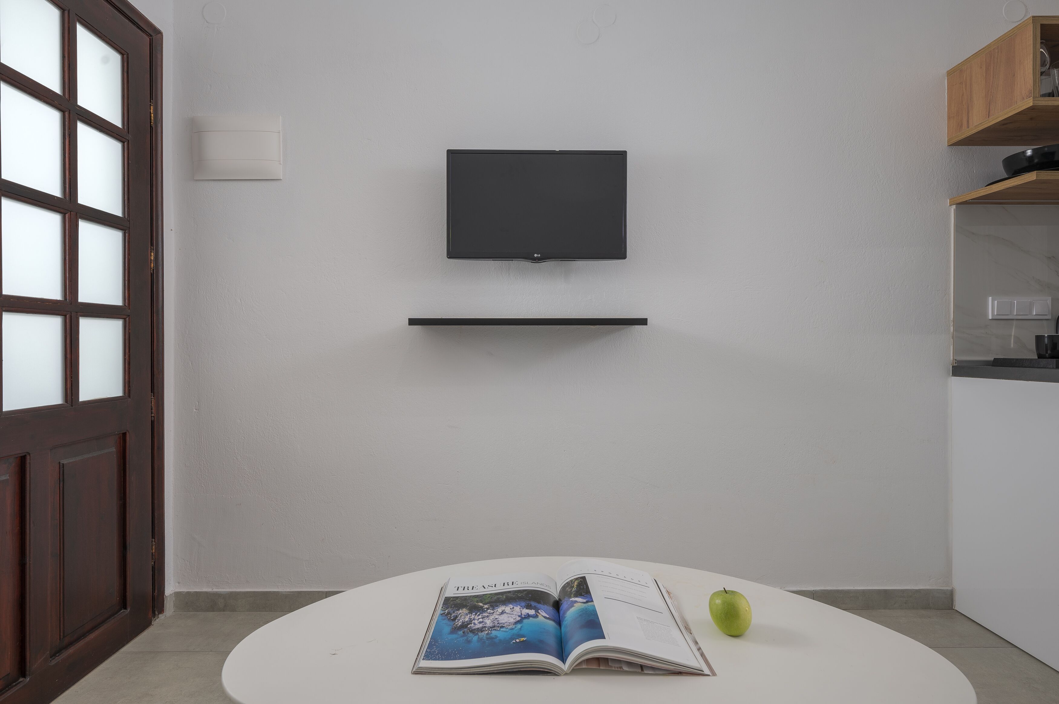 A modern room with a flat-screen TV mounted on the wall, a white table with a magazine and an apple, and a wooden cabinet with kitchen items.