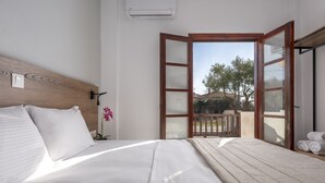 A bedroom with a wooden headboard, a large bed with white linens, a nightstand, a potted orchid, and an open door leading to a balcony with a view of trees and a building. - Eleon Suites & Apartments (Kassandra)