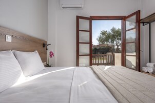 Quarto com cabeceira de madeira, cama grande com roupa de cama branca, mesa de cabeceira, orquídea em vaso e porta aberta para varanda com vista para árvores e um edifício.