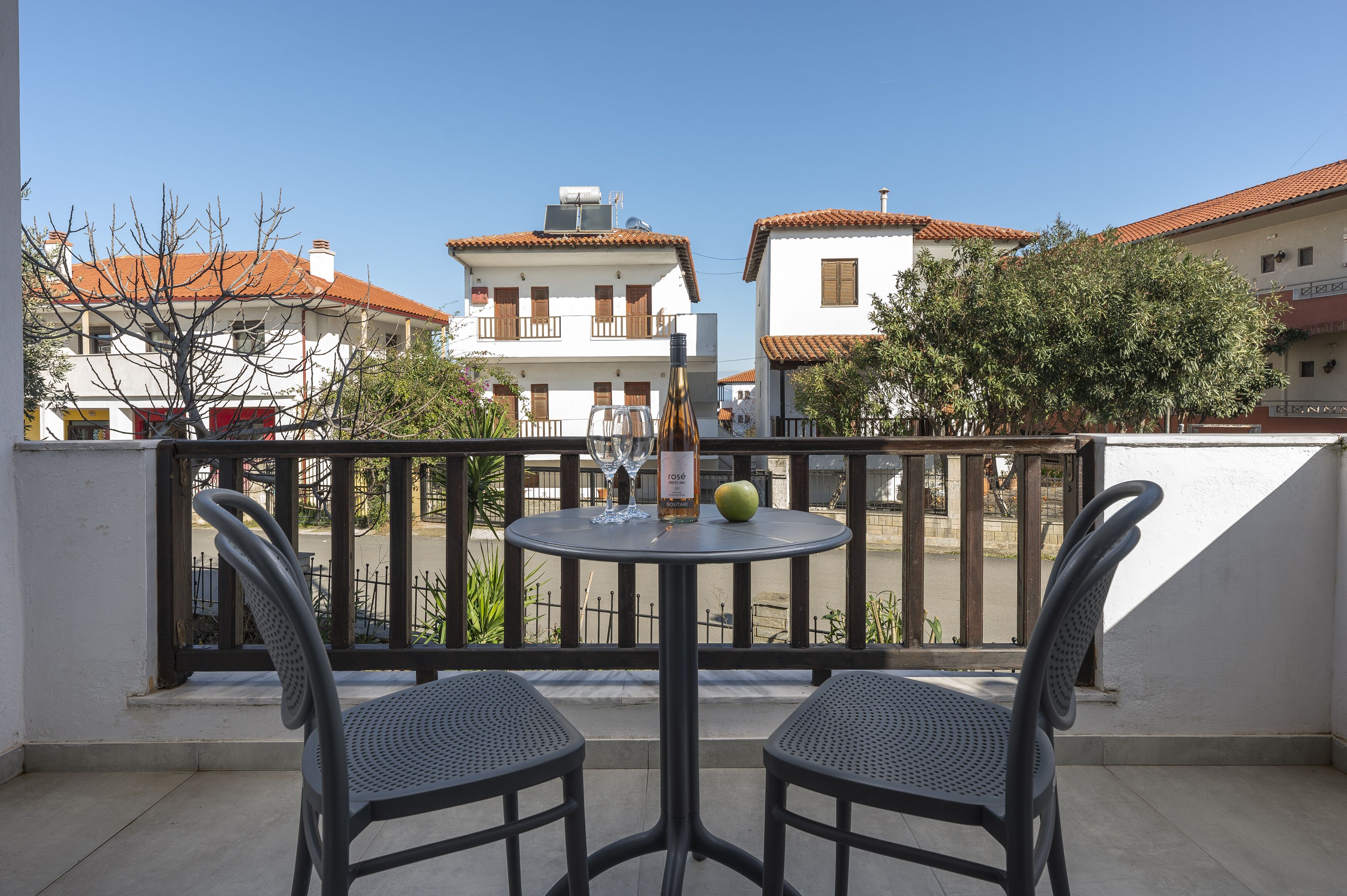 A balcony with two chairs and a table, overlooking a street with buildings and trees.