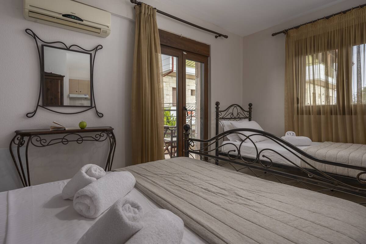 A hotel room with two beds, a bedside table with a book and an apple, a mirror, and a window with curtains.