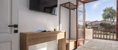 A bedroom with a bed, a wooden desk, a television mounted on the wall, and a sliding glass door leading to an outdoor balcony.