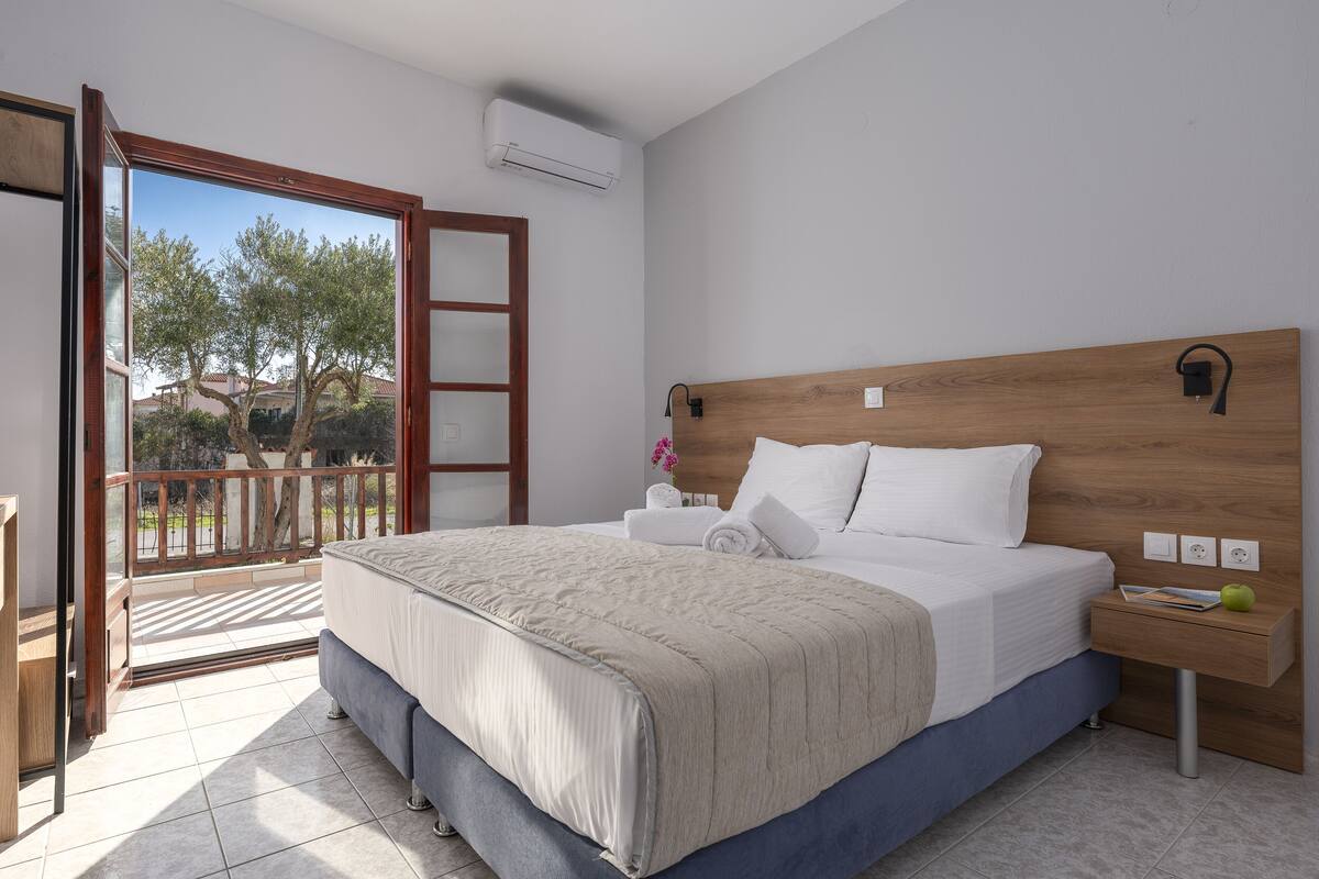 A bedroom with a wooden headboard, a large bed with a beige bedspread, a nightstand with a green apple, and a view of trees and a balcony.