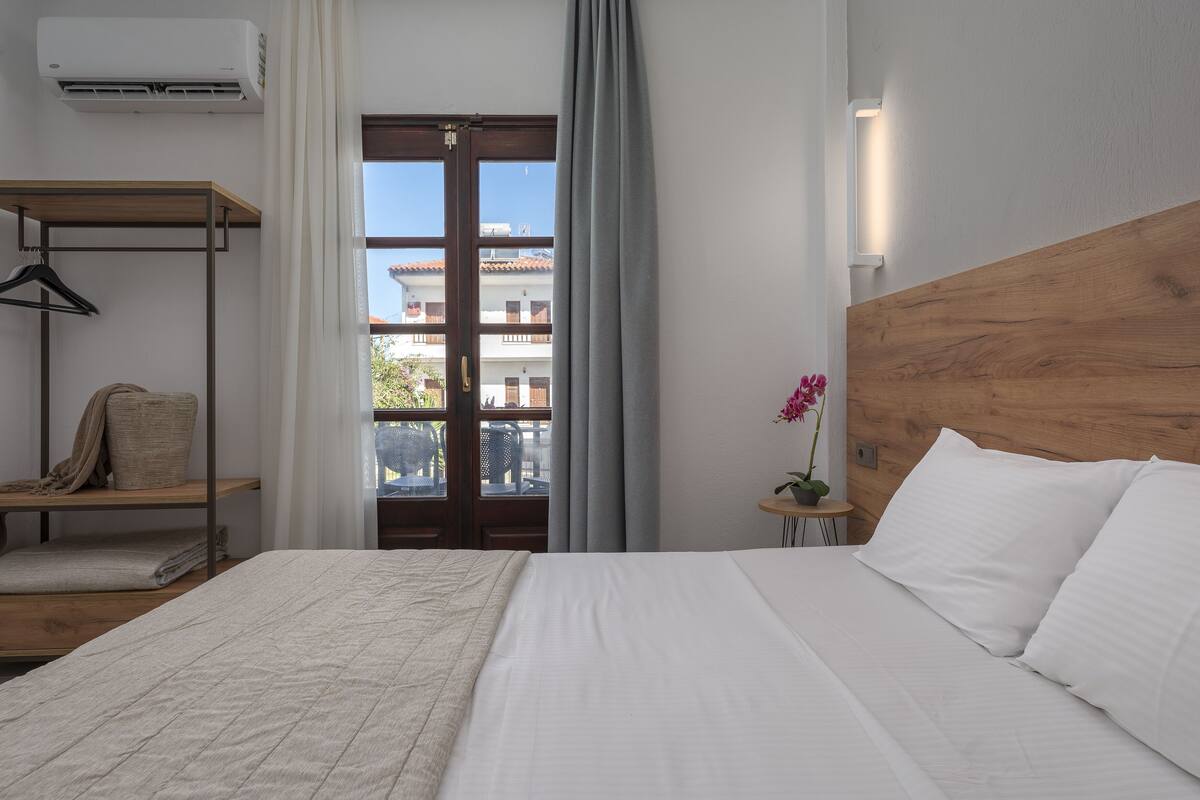 A hotel room with a bed, a wooden headboard, a window with curtains, a small table with a potted plant, and a hanging rack with a towel.