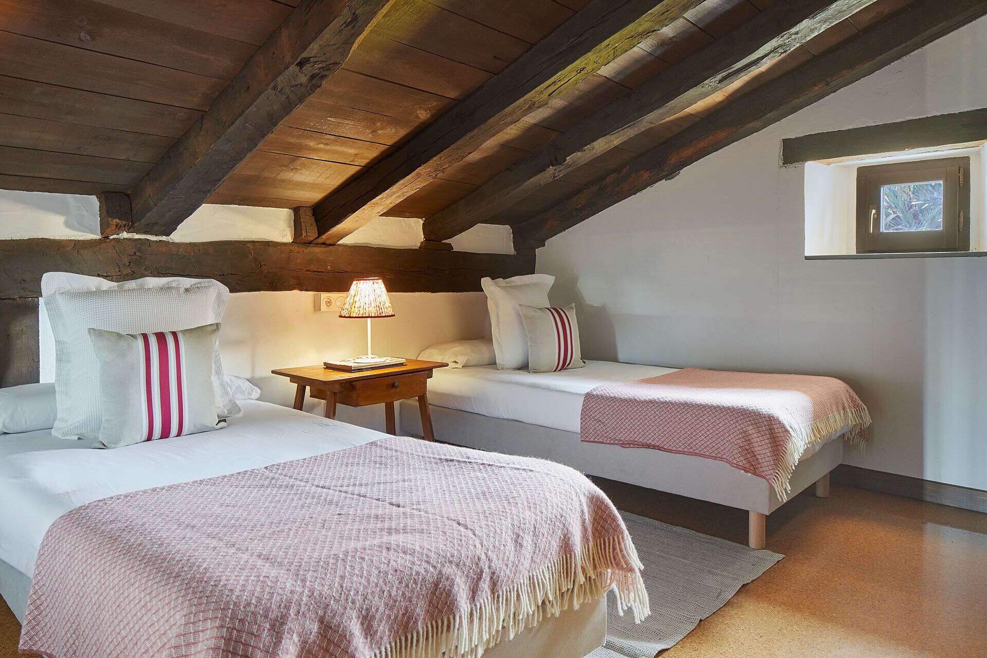 A cozy attic bedroom with two single beds, a nightstand with a lamp, and a small window.