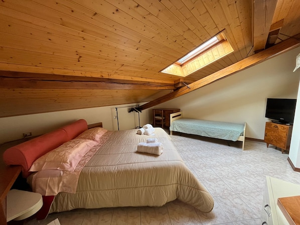 A bedroom with a sloped ceiling, a bed with a red pillow, a bench, and a television.