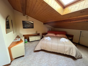 A bedroom with a sloped ceiling, a bed with a beige bedspread, a nightstand, a dresser, a mirror, and a framed picture on the wall.