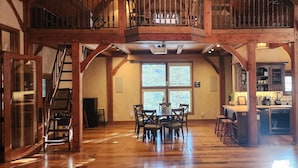 Une salle à manger spacieuse avec un plafond haut et des poutres en bois, une cuisine équipée d’appareils modernes et une grande fenêtre qui laisse entrer la lumière naturelle.
