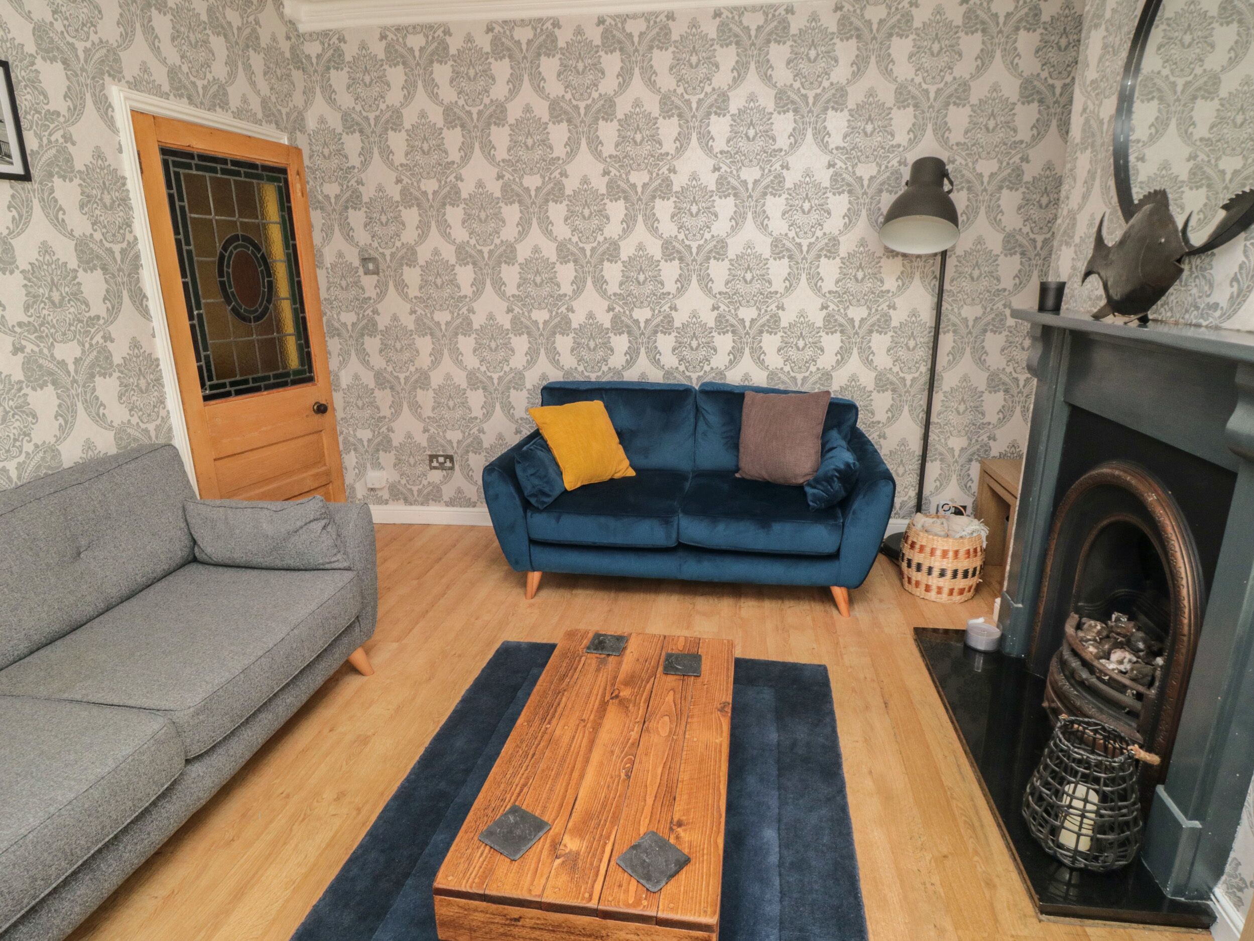 A living room with a grey sofa, a blue sofa with cushions, a wooden coffee table, a patterned wallpaper, a fireplace, and a lamp.