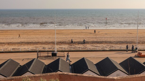 A beach with people walking, a no parking sign, and a row of buildings with sloped roofs. - Bay House (Bridlington)
