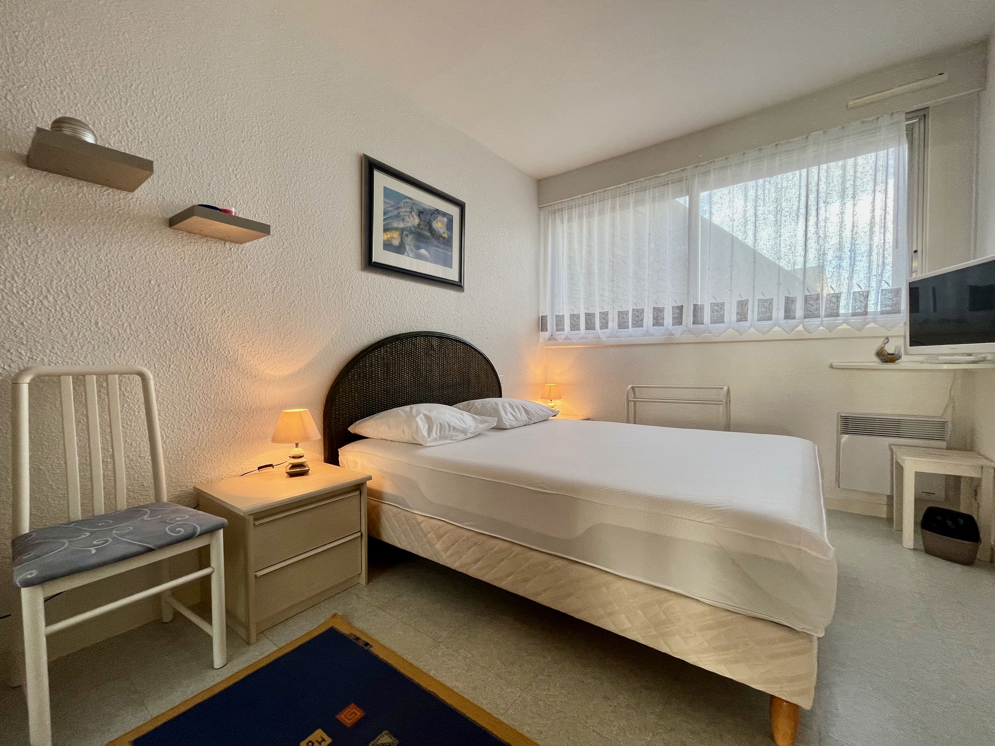 Een slaapkamer met een bed, nachtkastjes, een stoel, een televisie en een raam met gordijnen.