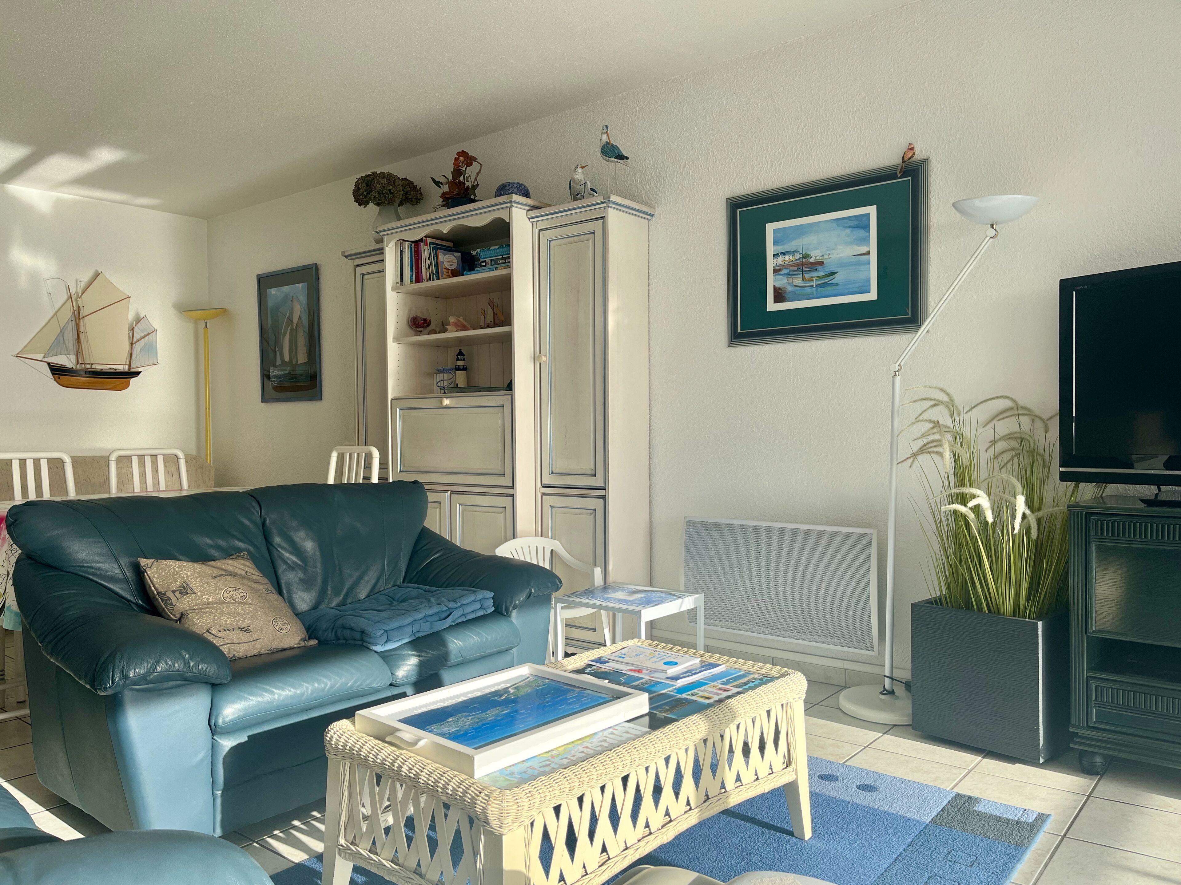 Un salon avec un canapé en cuir bleu, une petite table basse sur laquelle sont posés des magazines, une bibliothèque, un tableau encadré au mur, un téléviseur et un radiateur.