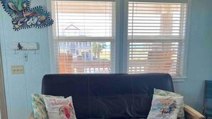 A room with a black leather sofa, two windows with blinds, a wall-mounted air conditioner, and a decorative piece above the sofa. - Cozy studio walking distance to the beach, Seas the Day Beach House (Cameron)