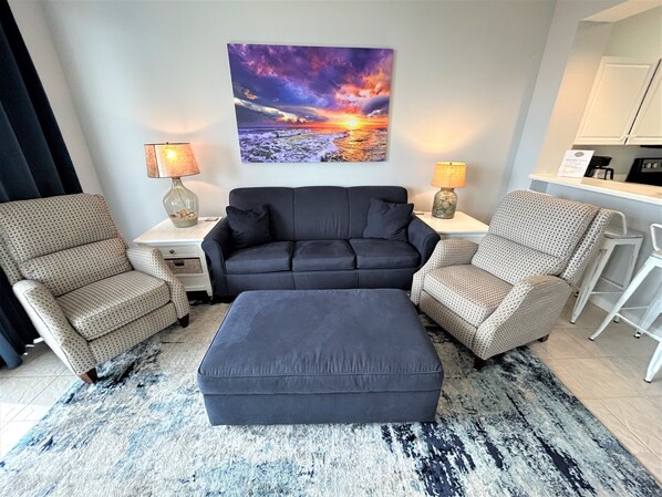 Un salon comprenant un canapé, deux fauteuils, une table basse et un tapis. Un tableau est accroché au mur, et une lampe est placée de chaque côté du canapé.