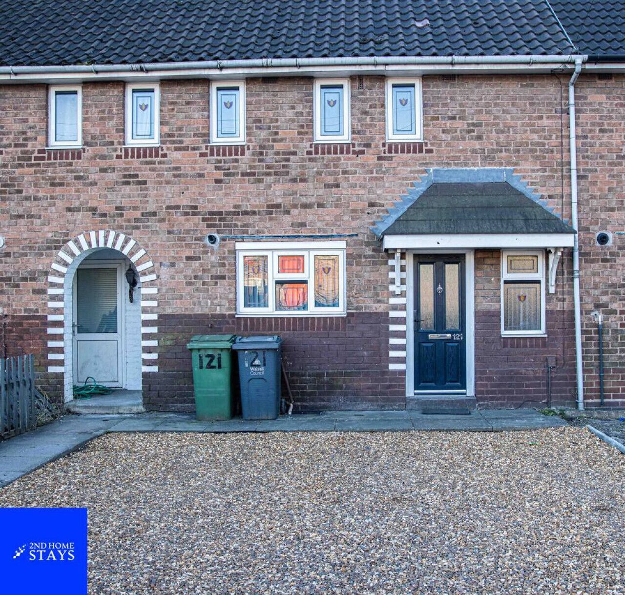 A brick building with a traditional design, featuring a central door with a number plate, a small window with a decorative frame, and a gravel driveway.