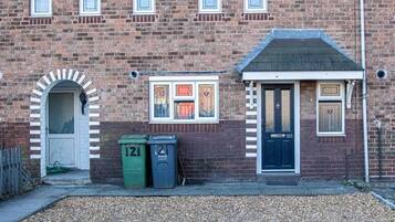 A brick building with a traditional design, featuring a central door with a number plate, a small window with a decorative frame, and a gravel driveway.