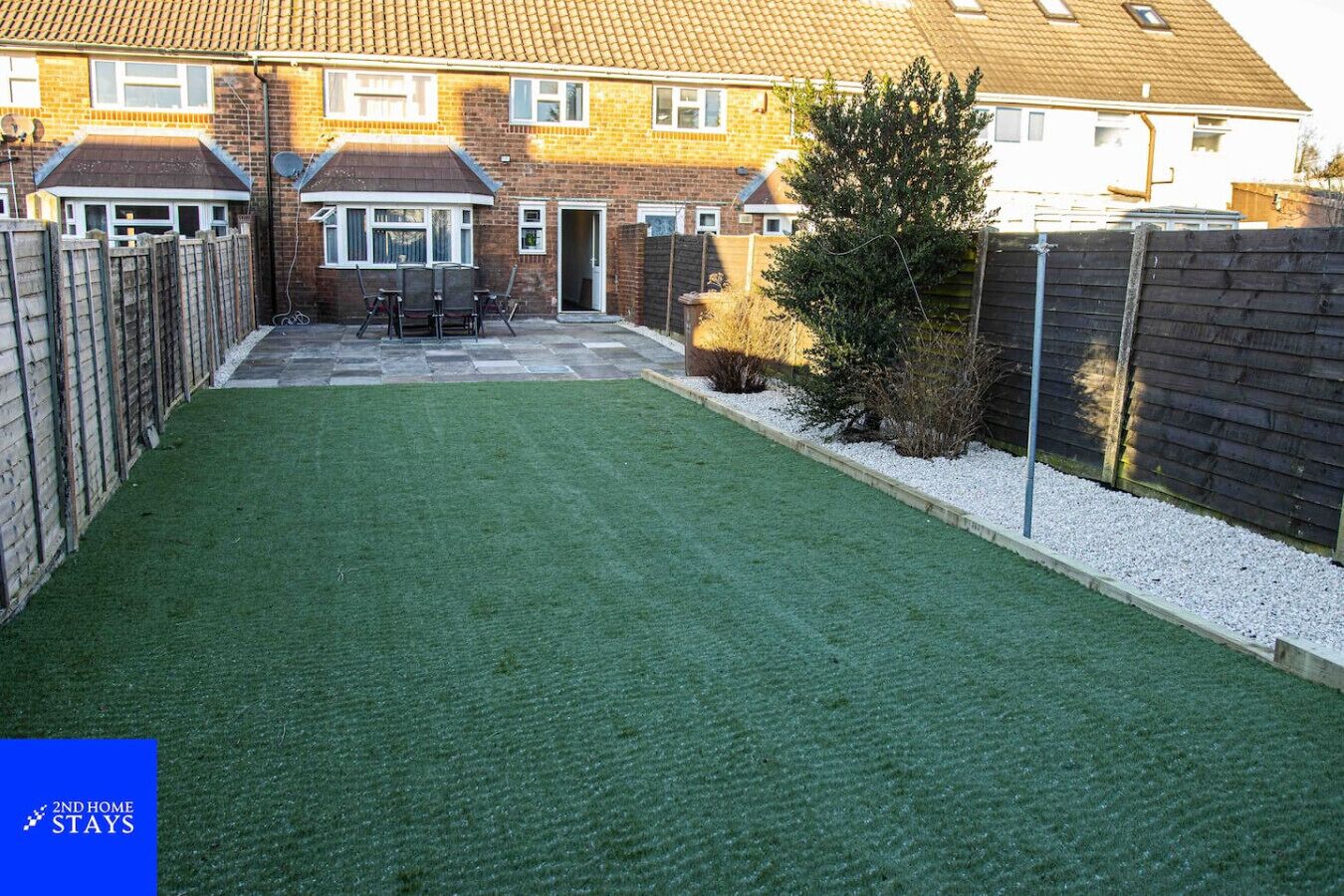 A residential backyard with artificial grass, a wooden fence, and a brick house.