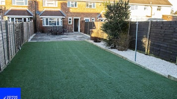 A residential backyard with artificial grass, a wooden fence, and a brick house.