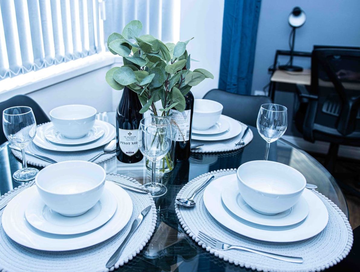 A dining table set with white plates, bowls, and cutlery, a bottle of wine, and a vase with green leaves.